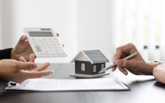 Un profesional ofrece asesoramiento durante la venta de un piso a un cliente, mostrando cálculos en una calculadora junto a una maqueta de casa y un contrato