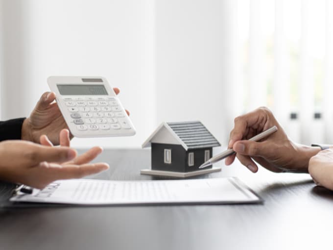 Un profesional ofrece asesoramiento durante la venta de un piso a un cliente, mostrando cálculos en una calculadora junto a una maqueta de casa y un contrato